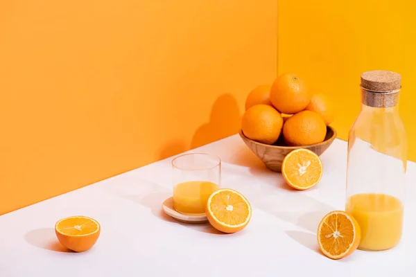 Suco de laranja fresco em vidro e garrafa perto de laranjas maduras em tigela na superfície branca no fundo laranja — Fotografia de Stock