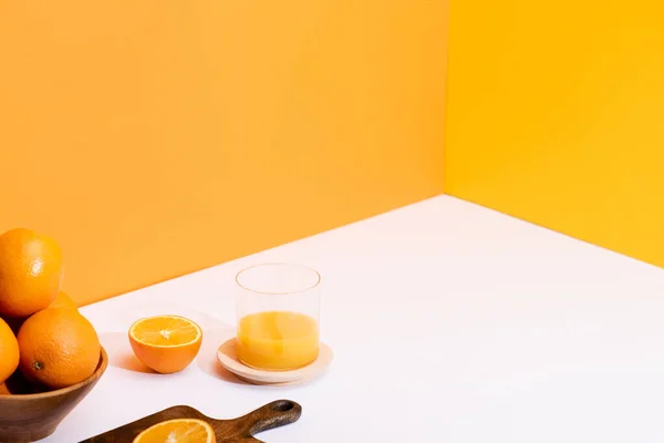 Frischer Orangensaft im Glas neben reifen Orangen in Schüssel und Holzschneidebrett auf weißer Oberfläche auf orangefarbenem Hintergrund — Stockfoto