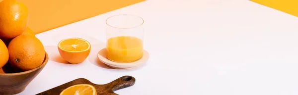 Fresh orange juice in glass near ripe oranges in bowl and wooden cutting board on white surface on orange background, panoramic shot — Stock Photo