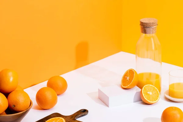 Zumo de naranja fresco en vidrio y botella cerca de naranjas maduras en tazón y tabla de cortar en la superficie blanca sobre fondo naranja - foto de stock