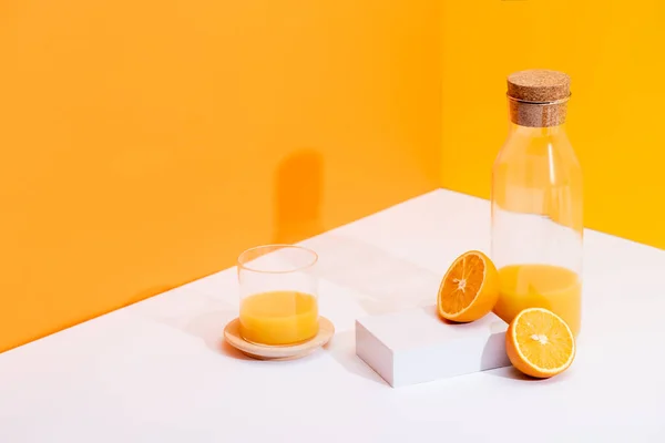 Zumo de naranja fresco en vidrio y botella cerca de naranjas maduras en la superficie blanca sobre fondo naranja - foto de stock