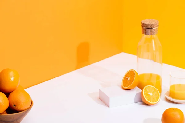 Zumo de naranja fresco en botella de vidrio cerca de naranjas maduras en un tazón sobre una superficie blanca sobre fondo naranja - foto de stock