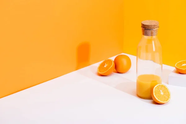 Zumo de naranja fresco en botella de vidrio cerca de naranjas maduras en la superficie blanca sobre fondo naranja - foto de stock