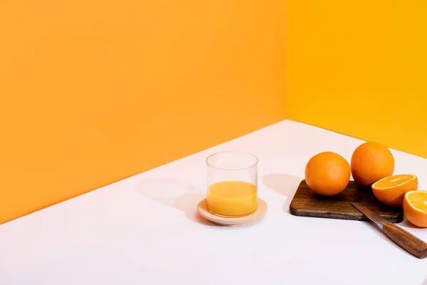 Frischer Orangensaft im Glas neben reifen Orangen auf Schneidebrett mit Messer auf weißer Oberfläche auf orangefarbenem Hintergrund — Stockfoto