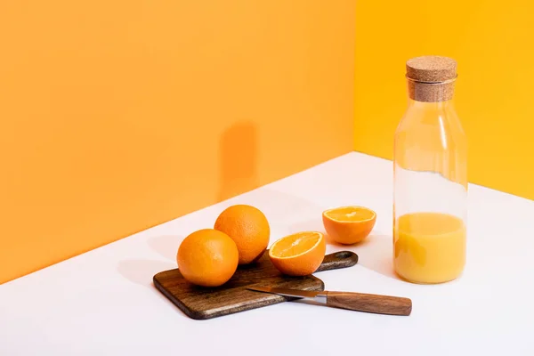 Suco de laranja fresco em garrafa de vidro perto de laranjas maduras na placa de corte com faca na superfície branca no fundo laranja — Fotografia de Stock