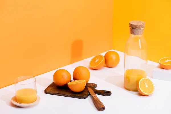 Suco de laranja fresco em vidro e garrafa perto de laranjas maduras na placa de corte com faca na superfície branca no fundo laranja — Fotografia de Stock