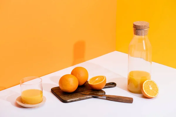 Suco de laranja fresco em vidro e garrafa perto de laranjas maduras na placa de corte com faca na superfície branca no fundo laranja — Fotografia de Stock