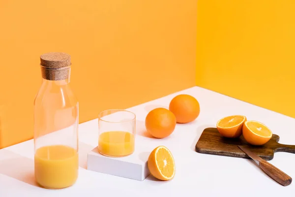 Frischer Orangensaft in Glas und Flasche nahe reifen Orangen auf Schneidebrett mit Messer auf weißer Oberfläche auf orangefarbenem Hintergrund — Stockfoto