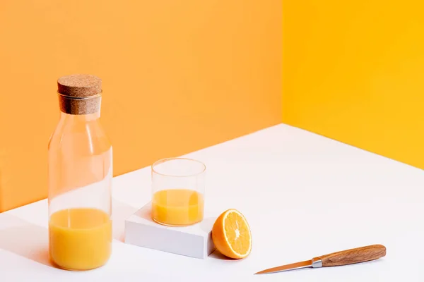 Suco de laranja fresco em vidro e garrafa perto de laranja madura e faca na superfície branca no fundo laranja — Fotografia de Stock