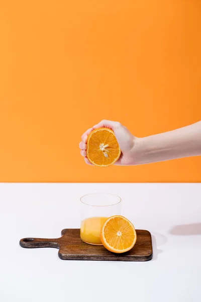 Vista recortada de la mujer exprimiendo jugo de naranja fresco en vidrio sobre tabla de cortar de madera en la superficie blanca aislada en naranja - foto de stock
