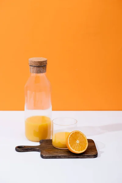 Fresh orange juice in glass and bottle near cut fruit on wooden cutting board on white surface isolated on orange — Stock Photo