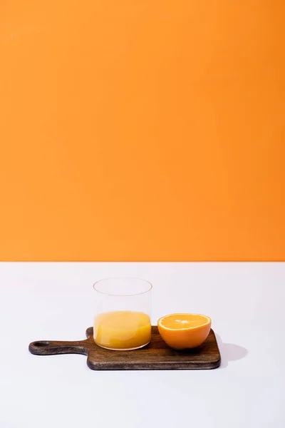 Jus d'orange frais en verre près de fruits coupés sur planche à découper en bois sur surface blanche isolé sur orange — Photo de stock