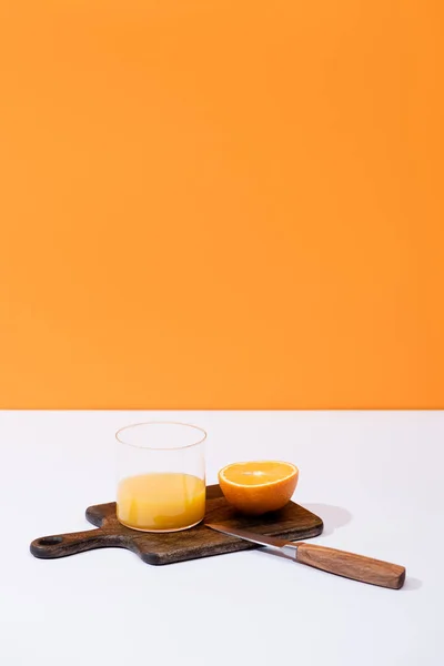 Fresh orange juice in glass near cut fruit on wooden cutting board with knife on white surface isolated on orange — Stock Photo