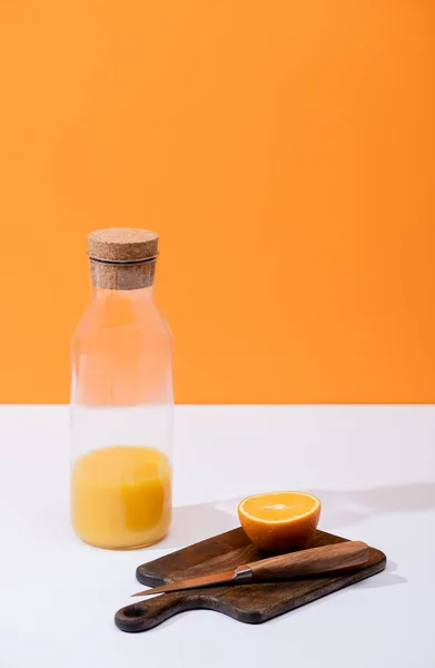 Fresh orange juice in glass bottle near cut fruit on wooden cutting board with knife on white surface isolated on orange — Stock Photo