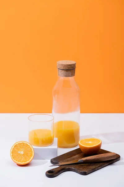 Fresh orange juice in glass and bottle near cut fruit on wooden cutting board with knife on white surface isolated on orange — Stock Photo