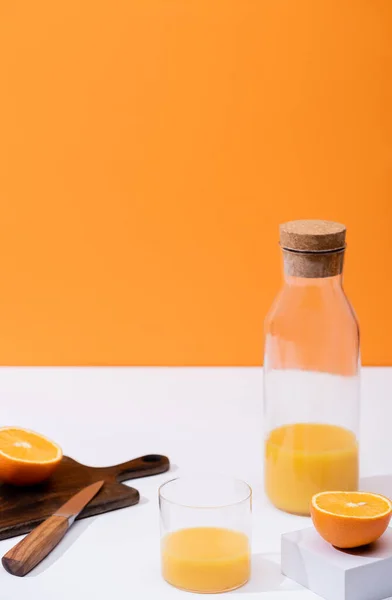 Fresh orange juice in glass and bottle near cut fruit on wooden cutting board with knife on white surface isolated on orange — Stock Photo