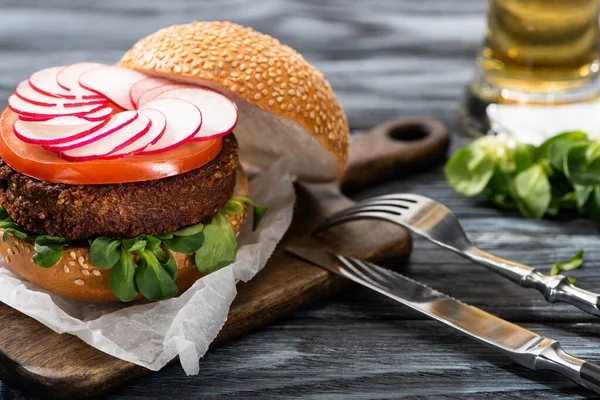 Selective focus of tasty vegan burger with radish and tomato served on cutting board with cutlery on wooden table — Stock Photo