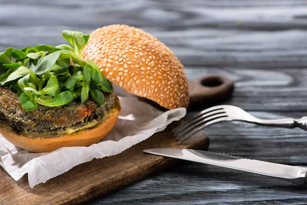 Hambúrguer vegan saboroso com microgreens servido na placa de corte com talheres na mesa de madeira — Fotografia de Stock