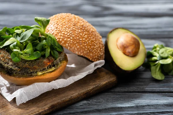 Вкусный веганский бургер с микрозелень подается на разделочной доске возле авокадо на деревянном столе — стоковое фото