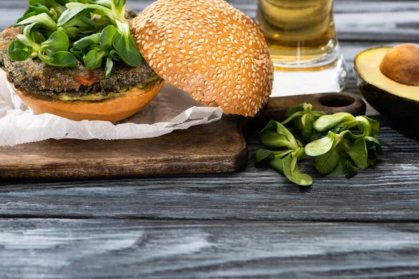 Hamburger végétalien savoureux avec microgreens servi sur planche à découper près de l'avocat et de la bière sur table en bois — Photo de stock