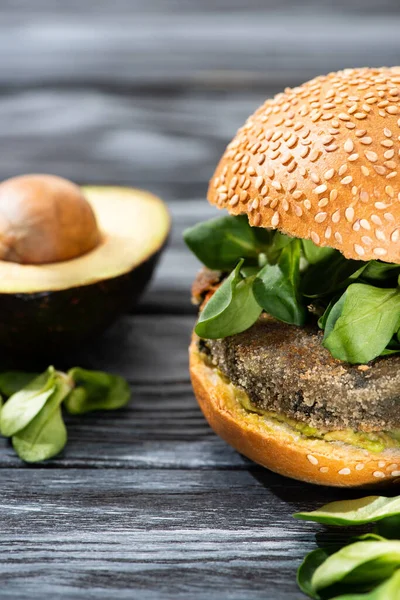 Избирательный фокус вкусного веганского бургера с микрозеленью подается на деревянном столе рядом с авокадо половина — стоковое фото