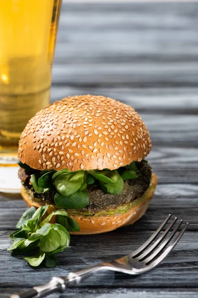 Selective focus of tasty vegan burger with microgreens served on wooden table with glass of beer and fork — Stock Photo