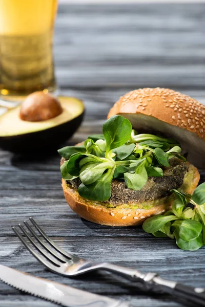Selective focus of tasty vegan burger with microgreens served on wooden table with avocado half, cutlery and beer — Stock Photo