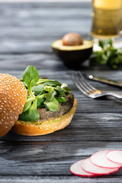 Selective focus of tasty vegan burger with microgreens served on wooden table with radish, avocado half, cutlery and beer — Stock Photo