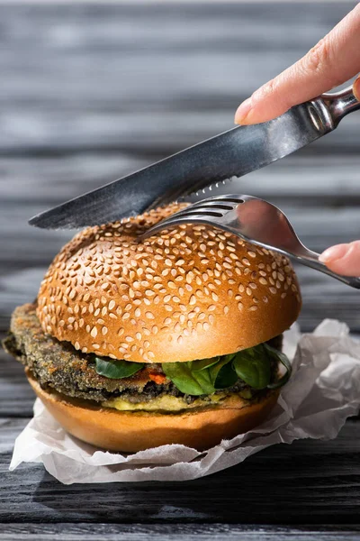 Cropped view of woman cutting tasty vegan burger with fork and knife — Stock Photo