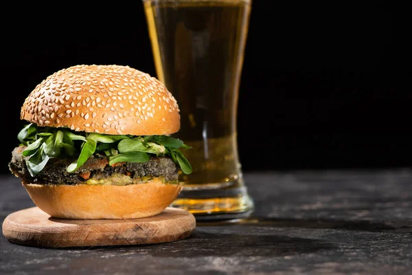 Messa a fuoco selettiva di gustoso hamburger vegan con microverdi serviti con birra su superficie testurizzata isolata su nero — Foto stock