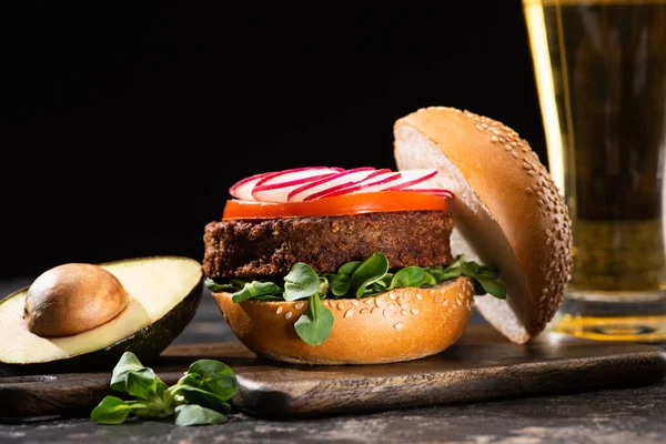 Selektiver Schwerpunkt des leckeren veganen Burgers mit Gemüse auf einem Holzbrett mit Avocado und Bier isoliert auf schwarz — Stockfoto