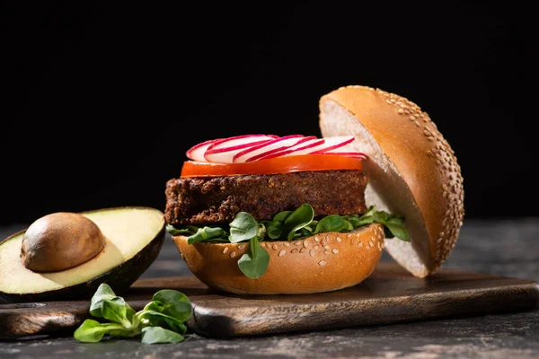 Tasty vegan burger with vegetables served on wooden cutting board with avocado isolated on black — Stock Photo