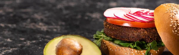 Sabrosa hamburguesa vegana con verduras servidas con aguacate mitad en superficie texturizada, cultivo panorámico - foto de stock