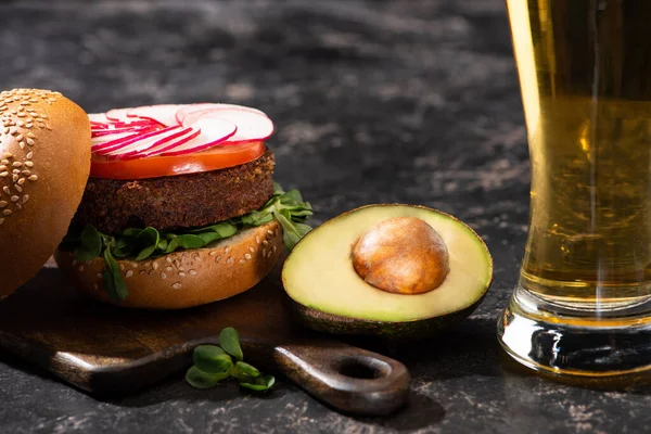 Sabrosa hamburguesa vegana con verduras servidas con mitad de aguacate y cerveza sobre tabla de cortar de madera en superficie texturizada - foto de stock