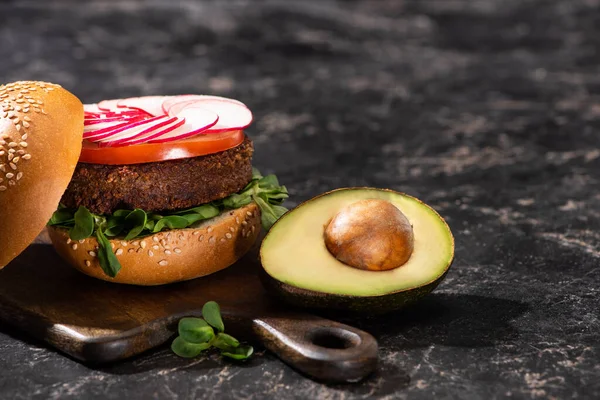 Tasty vegan burger with vegetables served with avocado half on wooden cutting board on textured surface — Stock Photo