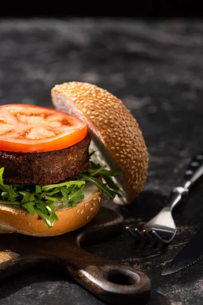 Sabrosa hamburguesa vegana con tomate y verduras servidas en tabla de cortar de madera con tenedor en superficie texturizada - foto de stock