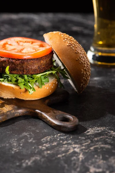 Selective focus of tasty vegan burger with tomato and greens served on wooden board on textured surface with beer — Stock Photo