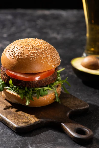 Selective focus of tasty vegan burger with tomato and greens served on wooden board on textured surface with beer and avocado — Stock Photo