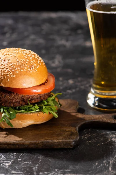 Foco seletivo de hambúrguer vegan saboroso com tomate e verduras servidos em tábua de madeira na superfície texturizada com cerveja — Fotografia de Stock