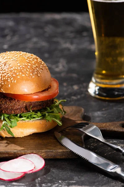 Selective focus of tasty vegan burger with tomato and greens served on wooden board on textured surface with beer and cutlery — Stock Photo