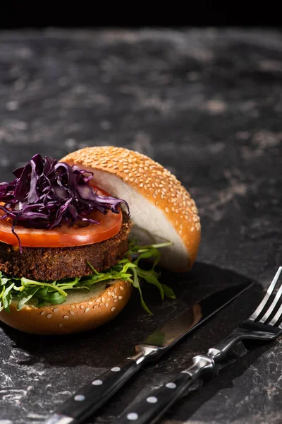 Tasty vegan burger with tomato, red cabbage and greens served on textured surface with cutlery — Stock Photo