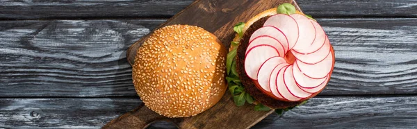 Top view of tasty vegan burger with microgreens, radish, tomato on cutting board served on wooden table, panoramic crop — Stock Photo
