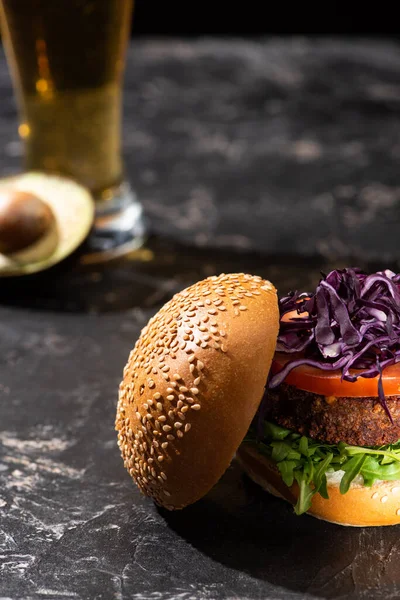 Selektiver Schwerpunkt des leckeren veganen Burgers mit Tomate, Rotkohl und Gemüse, serviert auf strukturierter Oberfläche mit Bier und Avocado — Stockfoto