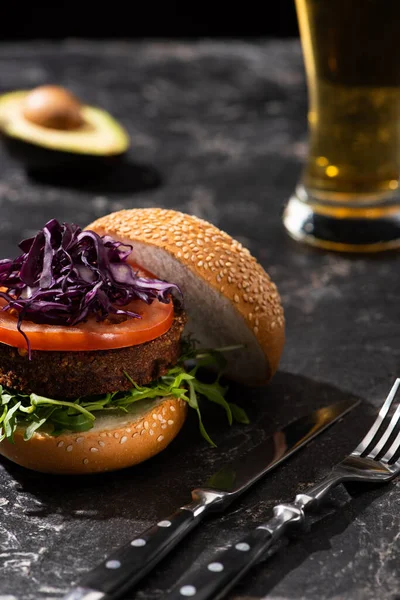 Selective focus of tasty vegan burger with tomato, red cabbage and greens served on textured surface with cutlery, beer and avocado — Stock Photo