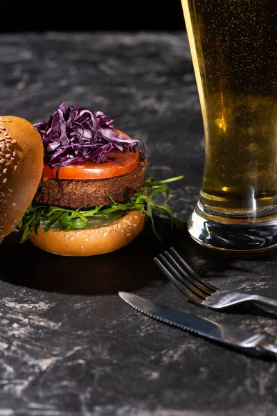 Sabrosa hamburguesa vegana con tomate, col roja y verduras servidas en superficie texturizada con cerveza y cubiertos - foto de stock