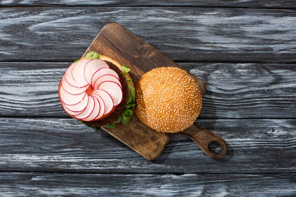 Top view of tasty vegan burger with microgreens, radish, tomato on cutting board served on wooden table — Stock Photo