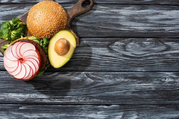 Top view of tasty vegan burger with microgreens, radish, tomato and avocado on cutting board served on wooden table — Stock Photo