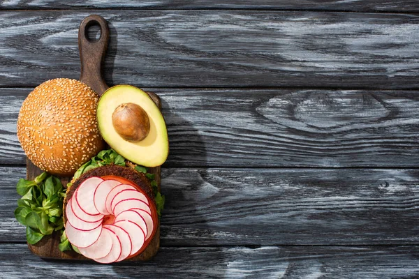 Top view of tasty vegan burger with microgreens, radish, tomato and avocado on cutting board served on wooden table — Stock Photo