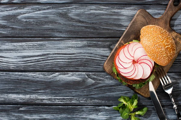 Top view of tasty vegan burger with microgreens, radish, tomato on cutting board with cutlery served on wooden table — Stock Photo