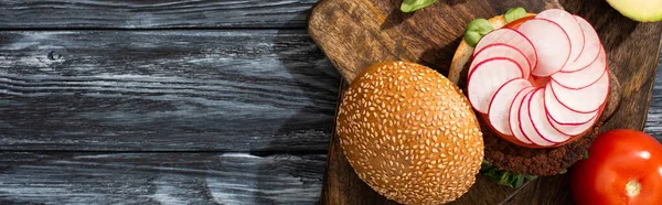 Top view of tasty vegan burger with microgreens, radish, tomato on cutting board served on wooden table, panoramic crop — Stock Photo